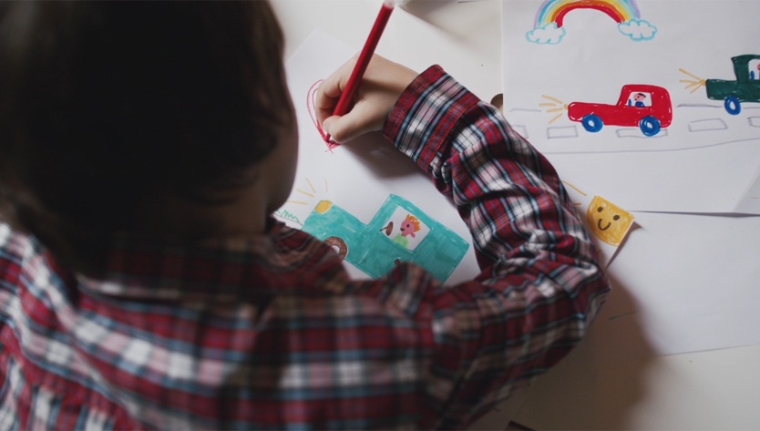 A child in a plaid shirt draws colorful cars with a red pencil. Nearby is a smiling face and a rainbow drawing, conveying creativity and joy.