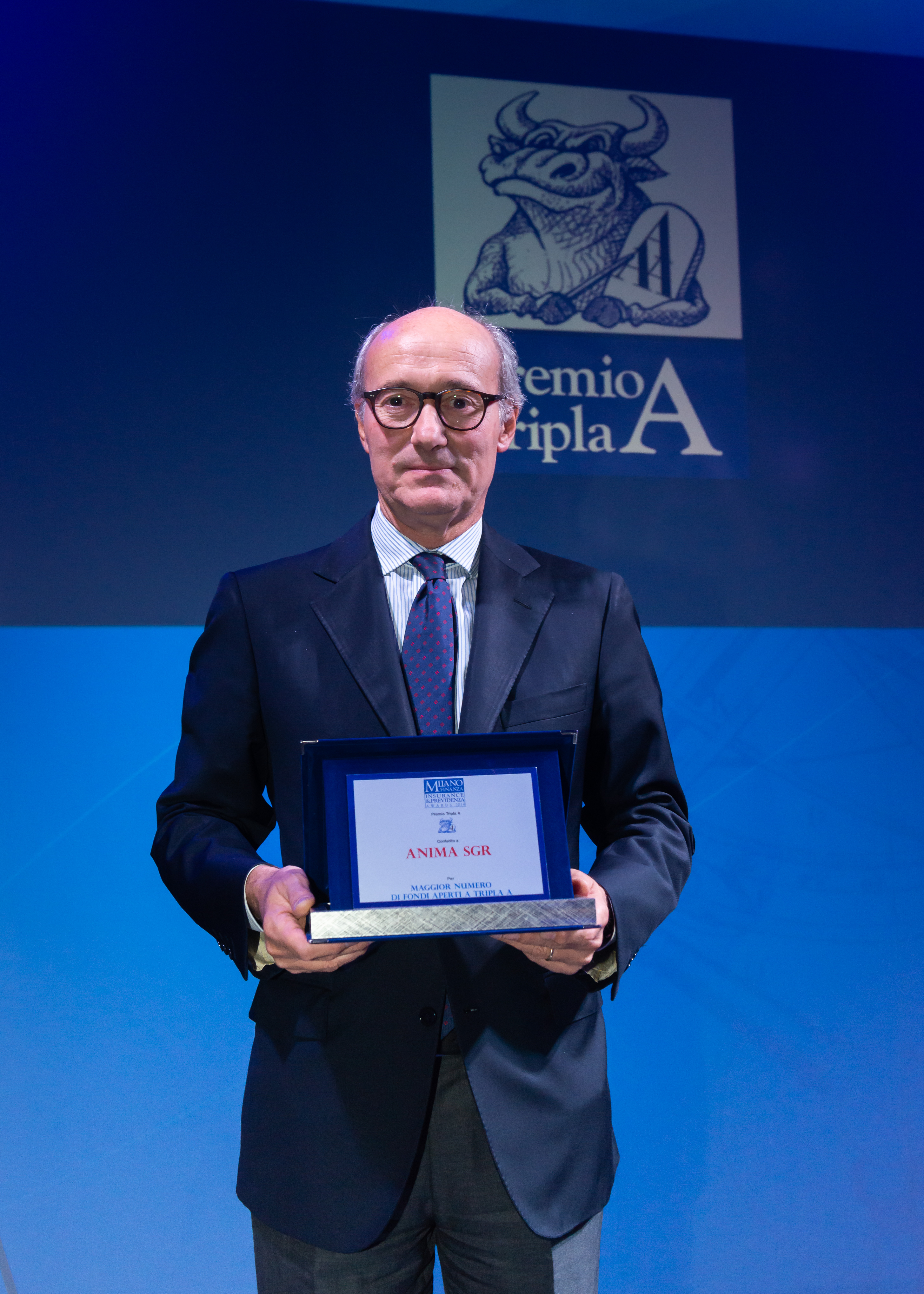 Un uomo in abito formale con occhiali tiene in mano una targa durante la cerimonia di premiazione del 
