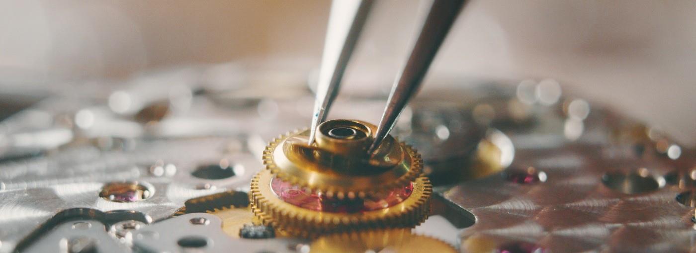 Primo piano di un orologiaio che inserisce un piccolo ingranaggio dorato nel meccanismo di un orologio usando delle pinzette.