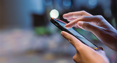 Primo piano di mani che utilizzano uno smartphone in condizioni di scarsa illuminazione, con sfondo sfocato. La scena trasmette concentrazione e connettività in un ambiente moderno.