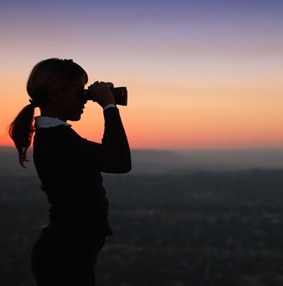 Una persona è in controluce contro un tramonto colorato, tiene in mano una fotocamera e guarda in lontananza.