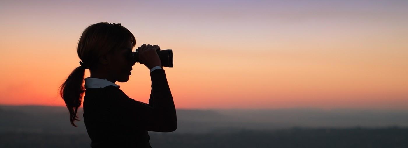 Una persona è in controluce contro un tramonto colorato, tiene in mano una fotocamera e guarda in lontananza.