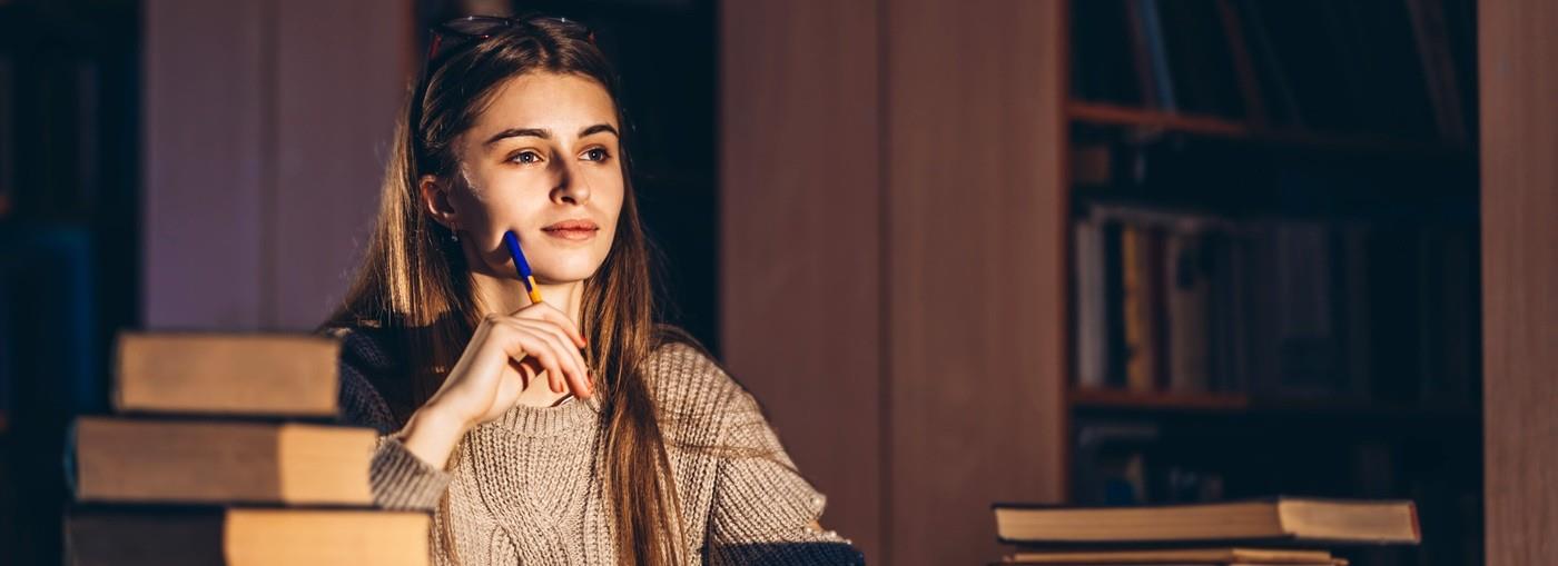 Una ragazza che studia fino a tarda ora a una scrivania circondata da pile di libri in una stanza poco illuminata.