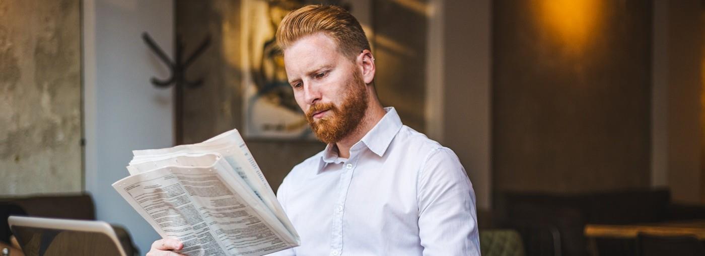 Un uomo con una camicia bianca è seduto a un tavolo, legge un giornale e tiene in mano una tazza di caffè, con un computer porta