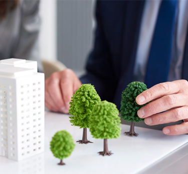 Modello di un paesaggio urbano con edifici bianchi e alberi verdi su un tavolo. Due mani, una delle quali indossa un abito, stanno sistemando il modello, suggerendo un'attività di progettazione o pianificazione.