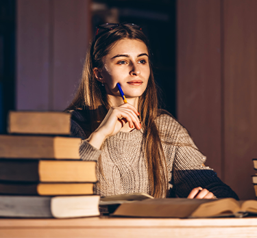Una donna dai capelli lunghi è seduta a un tavolo di legno circondata da libri, immersa nei suoi pensieri con una penna in mano. Indossa un maglione comodo ed è avvolta da una luce calda.